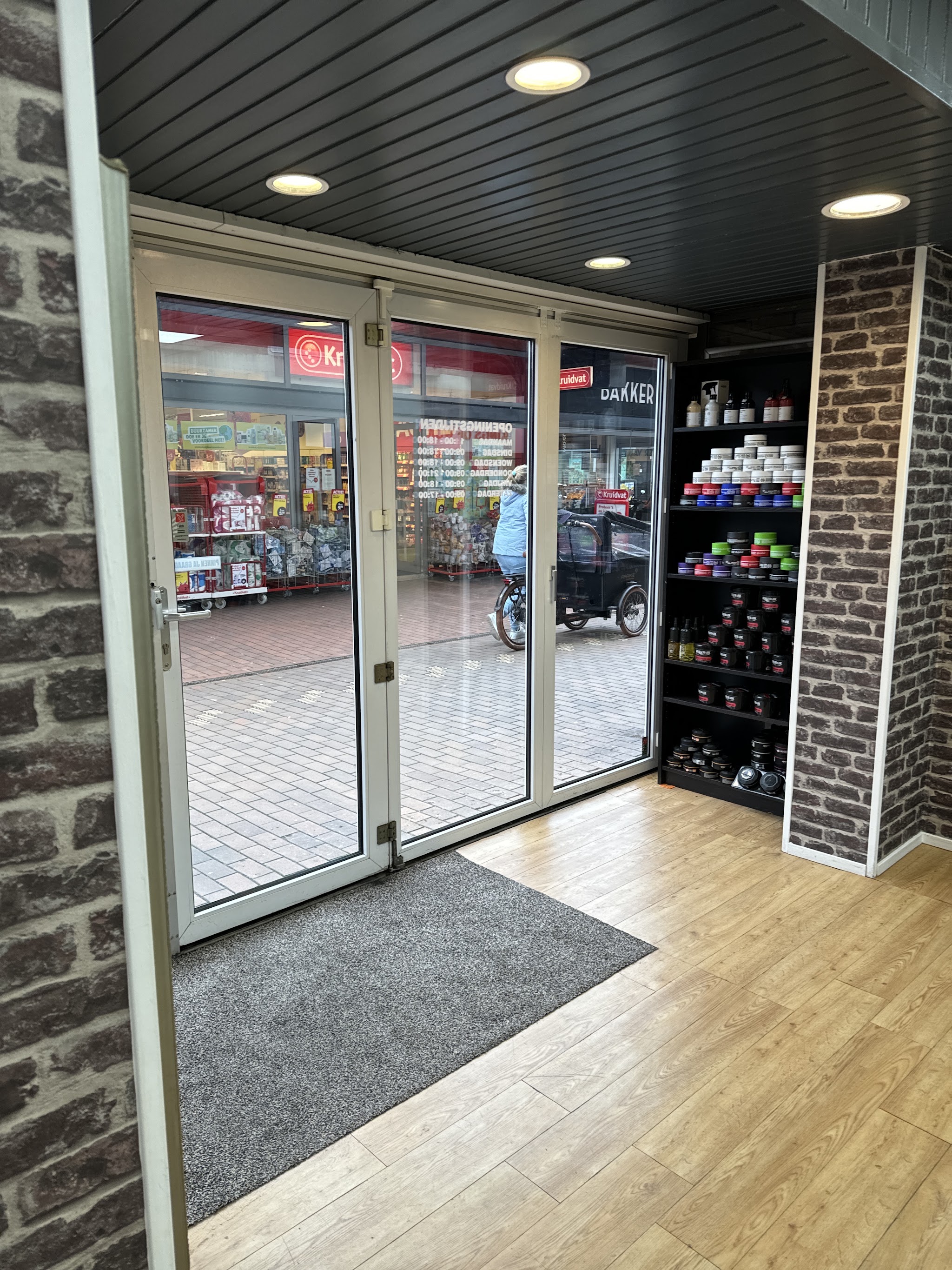 De ingang van Kapsalon HAKAR van binnenuit, met de glazen deuren naar het plein en een rek met haarproducten